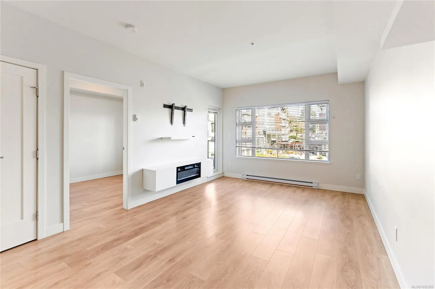 Bright, empty living room with light wood floors, white walls, and a large window with blinds. A modern fireplace and TV mount are on the wall.