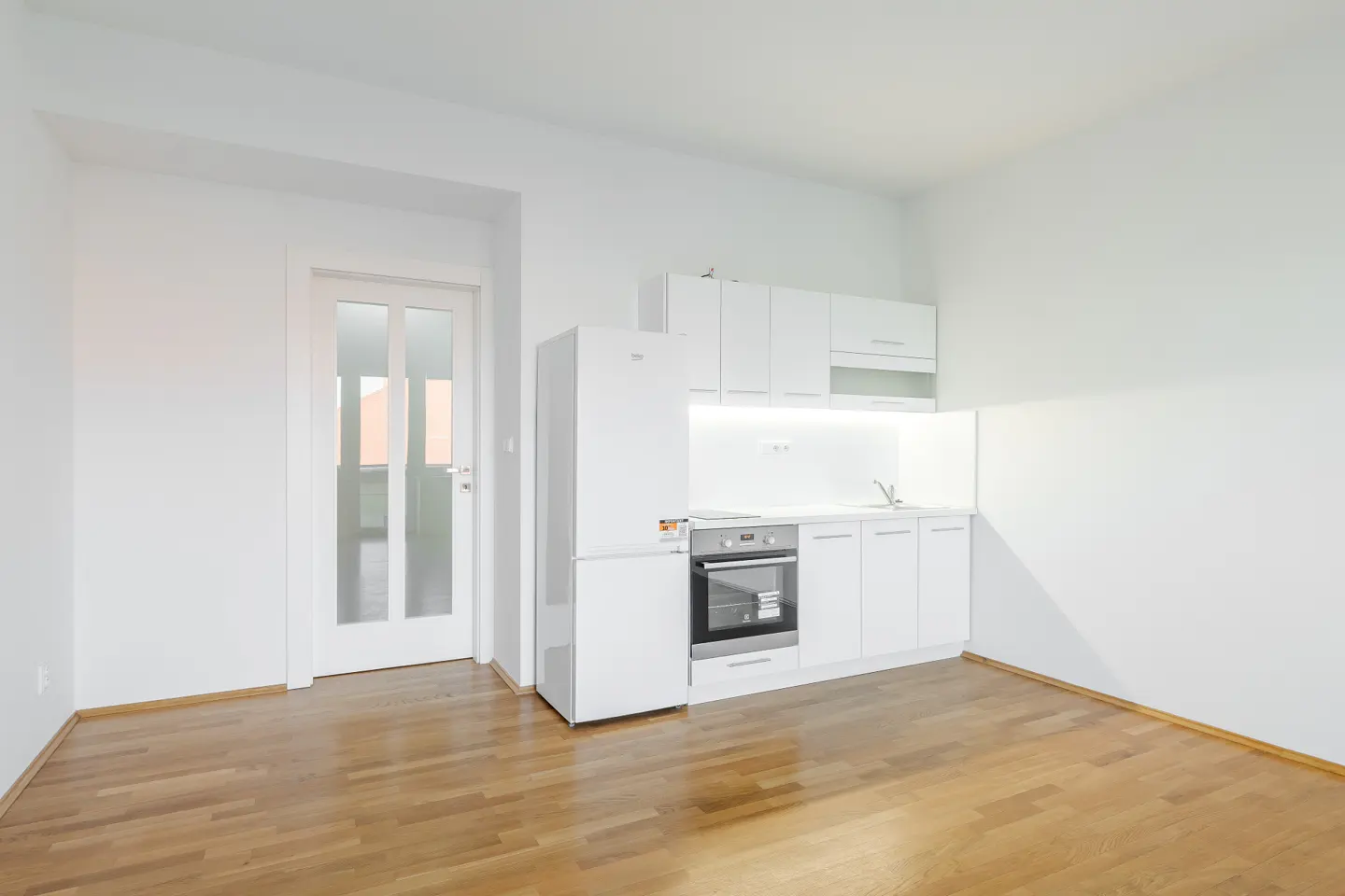 Bright, empty kitchen with white cabinets, fridge, oven, and wood floor. A glass door is on the left.