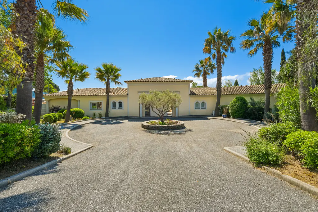 Exterior view of a one-story beige house with a gravel driveway, palm trees, and a blue sky.