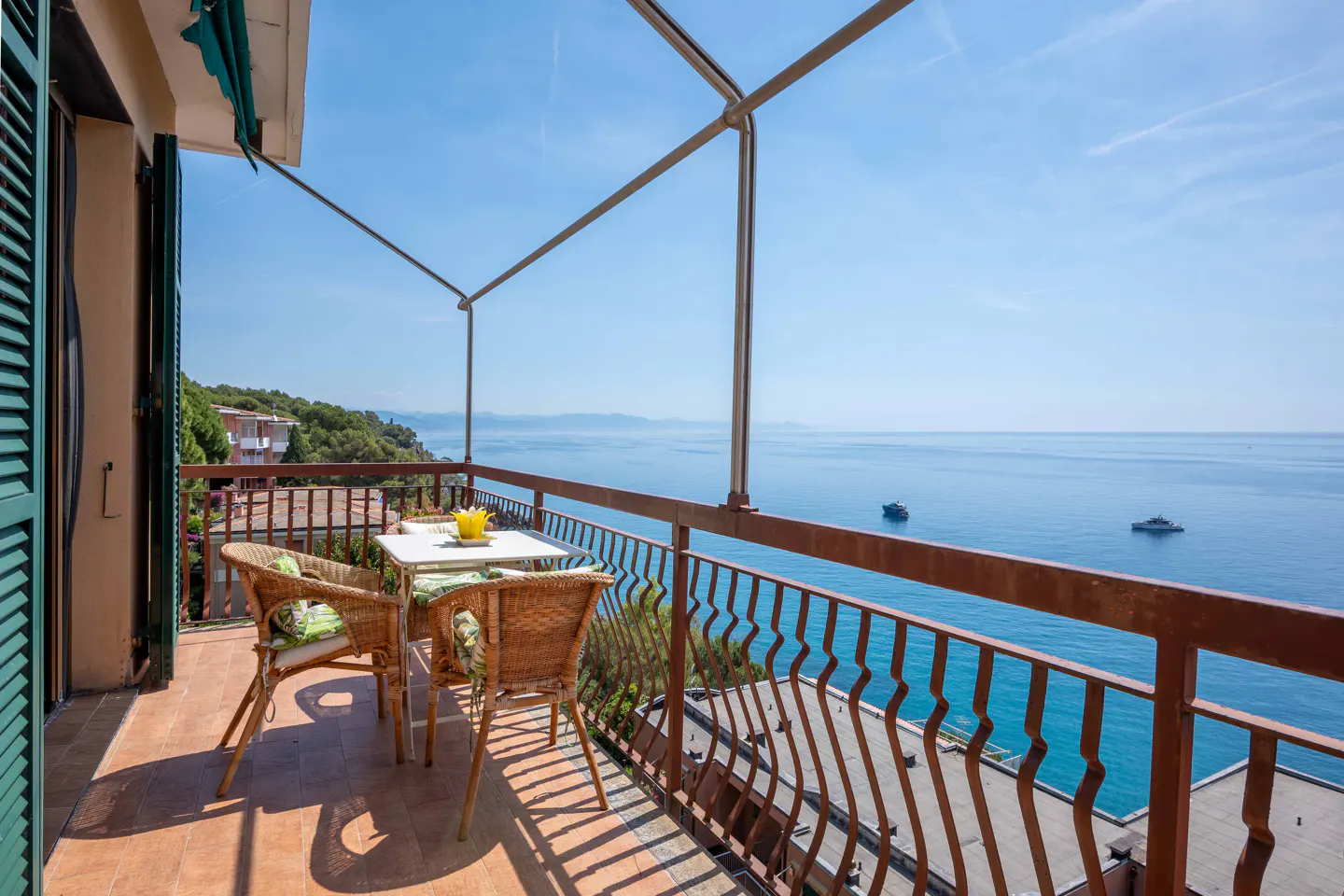 Balcony with wicker chairs and a small table overlooking a calm blue sea with boats under a clear sky.
