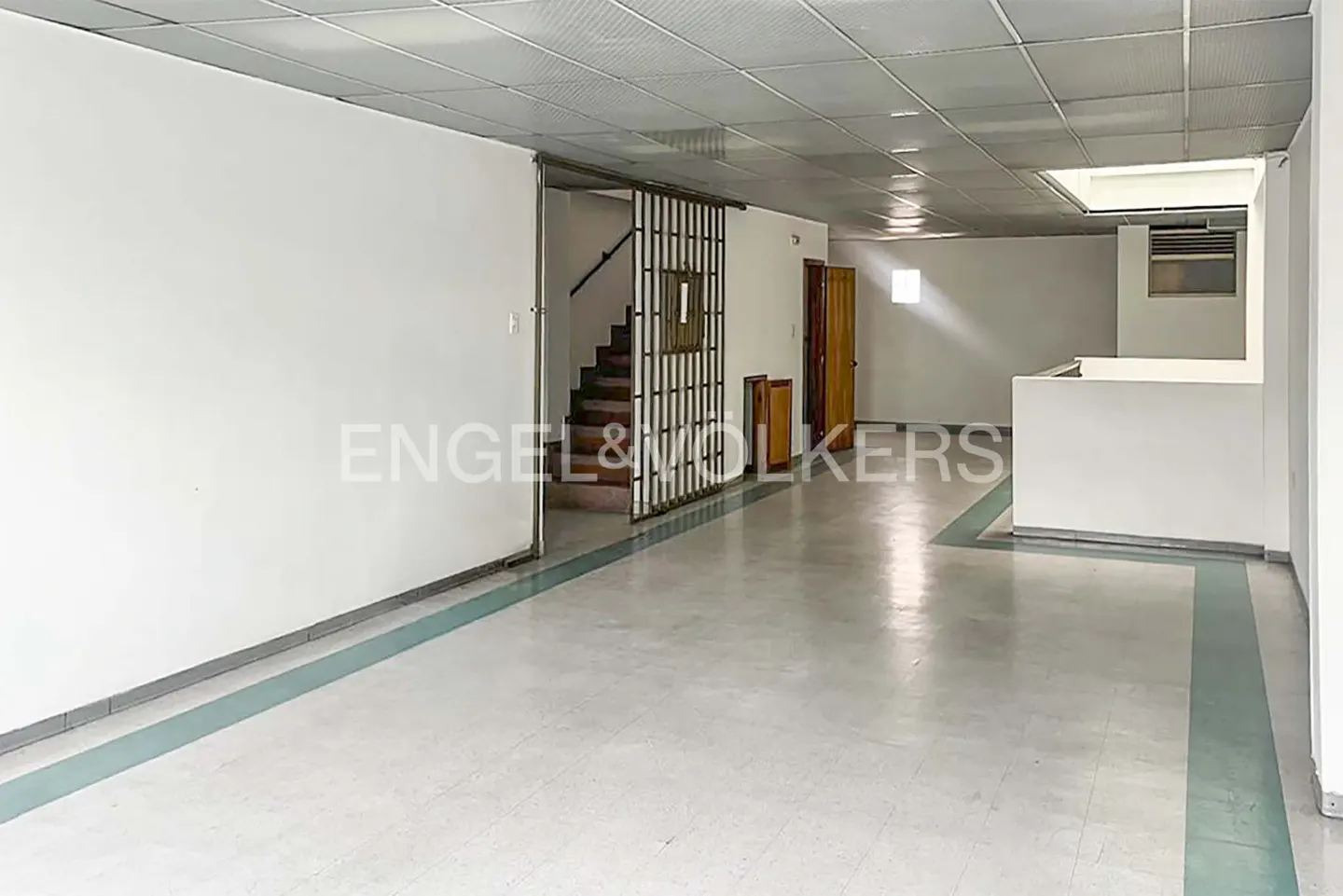 An empty room with white walls, a tiled floor with green accents, and a metal grid ceiling. Stairs and a door are visible in the background.