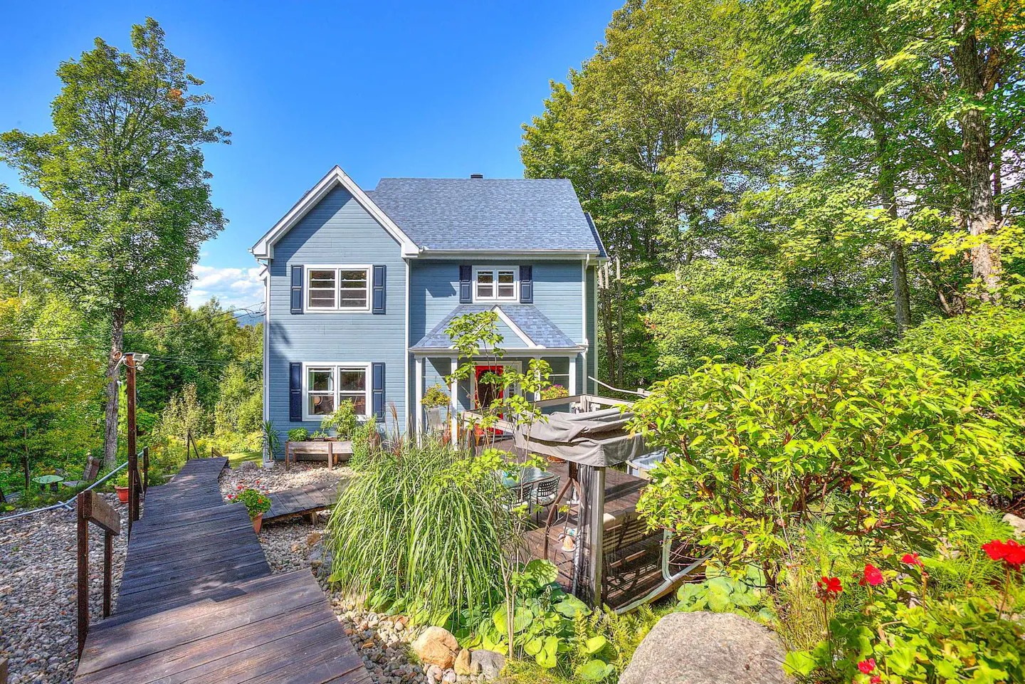 Two-story blue house with a red door, surrounded by lush green trees and a wooden walkway.
