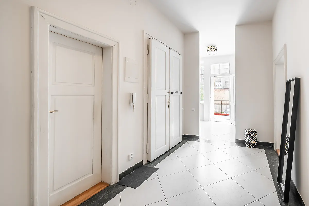 Bright hallway with white walls and tiled floor. A white door is on the left, and a double closet is in the center. A mirror leans against the wall on the right.