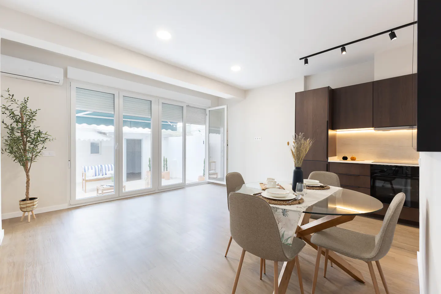 Bright, modern dining area with glass table, four chairs, and dark wood kitchen cabinets. Large glass doors lead to a patio with a blue and white striped bench.