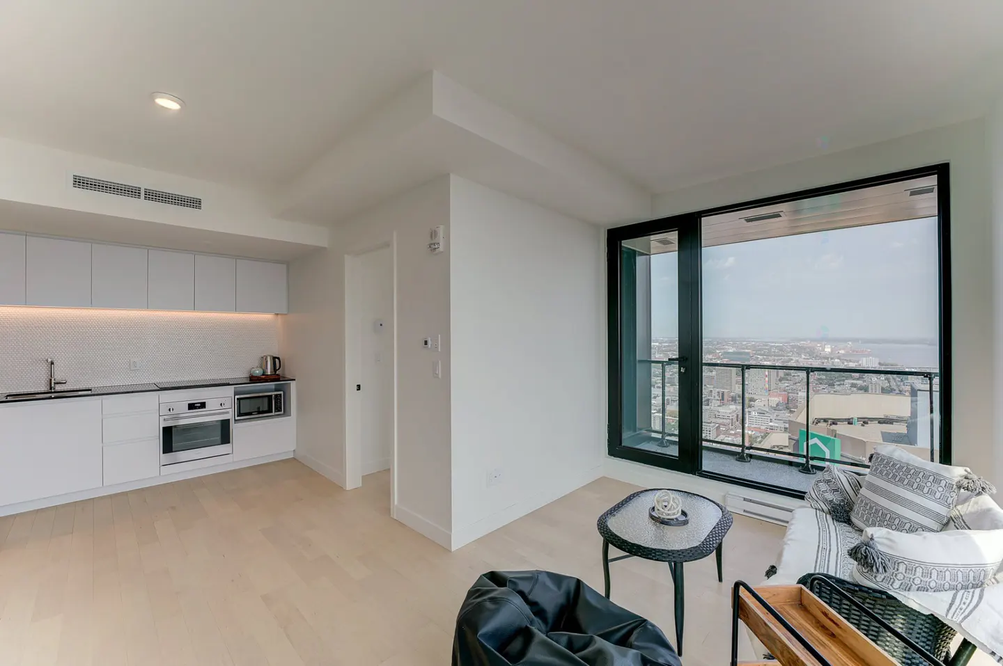 Bright, modern condo interior with white kitchen, light wood floors, and a large window with a city view. A black beanbag chair sits in the foreground.