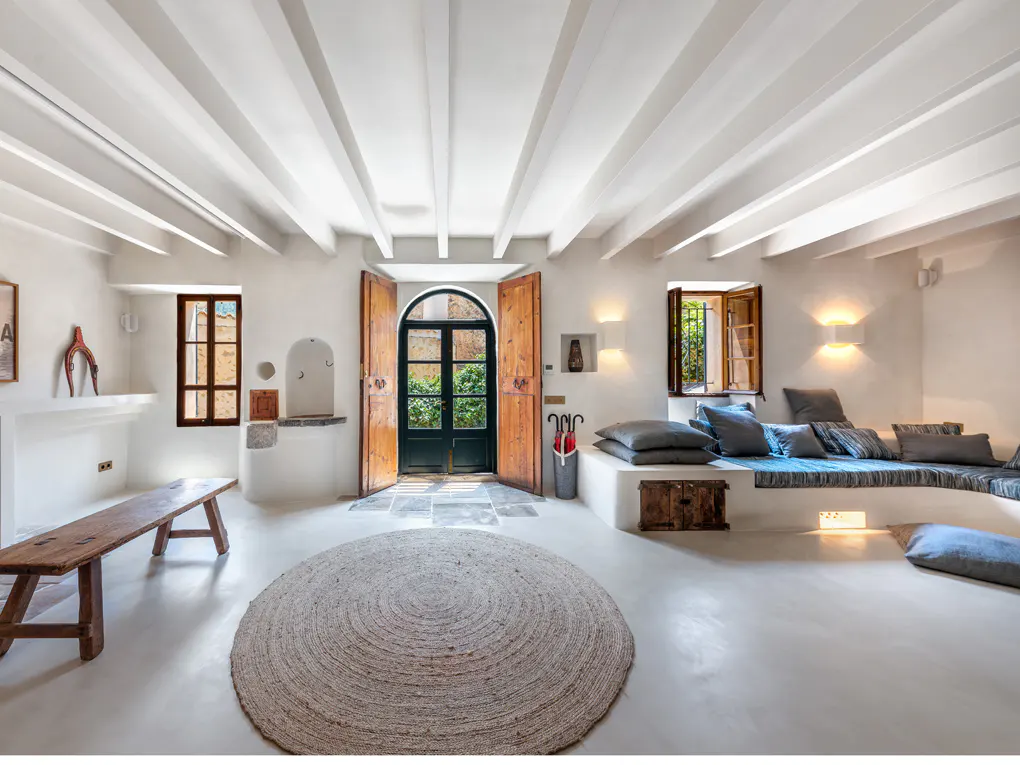Bright, airy living room with white walls, beamed ceiling, and a large round rug. Wooden bench, built-in seating with pillows, and open wooden doors to a garden.