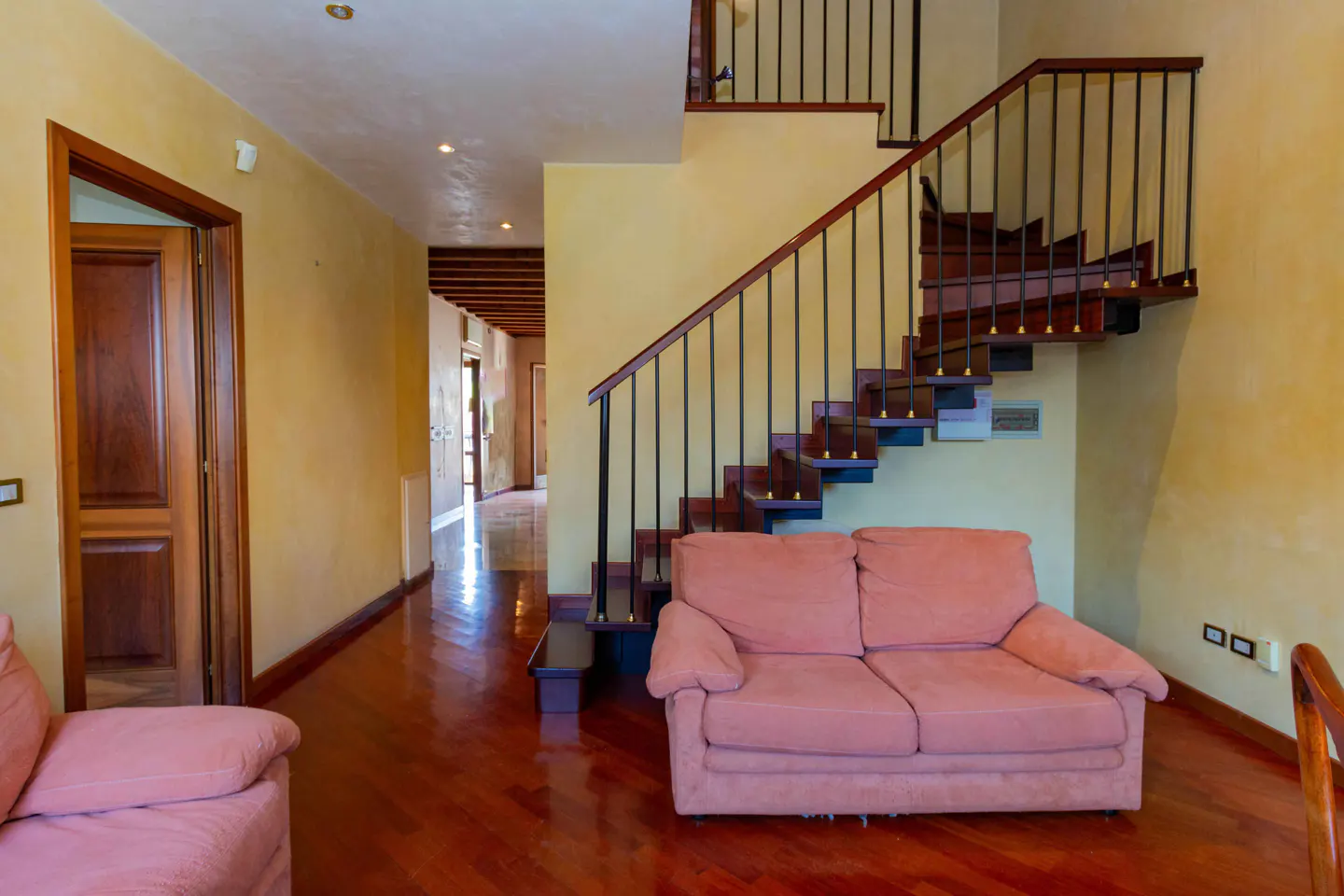 Interior view of a home with a pink sofa, wooden staircase with black railings, and a hallway with wooden floors.