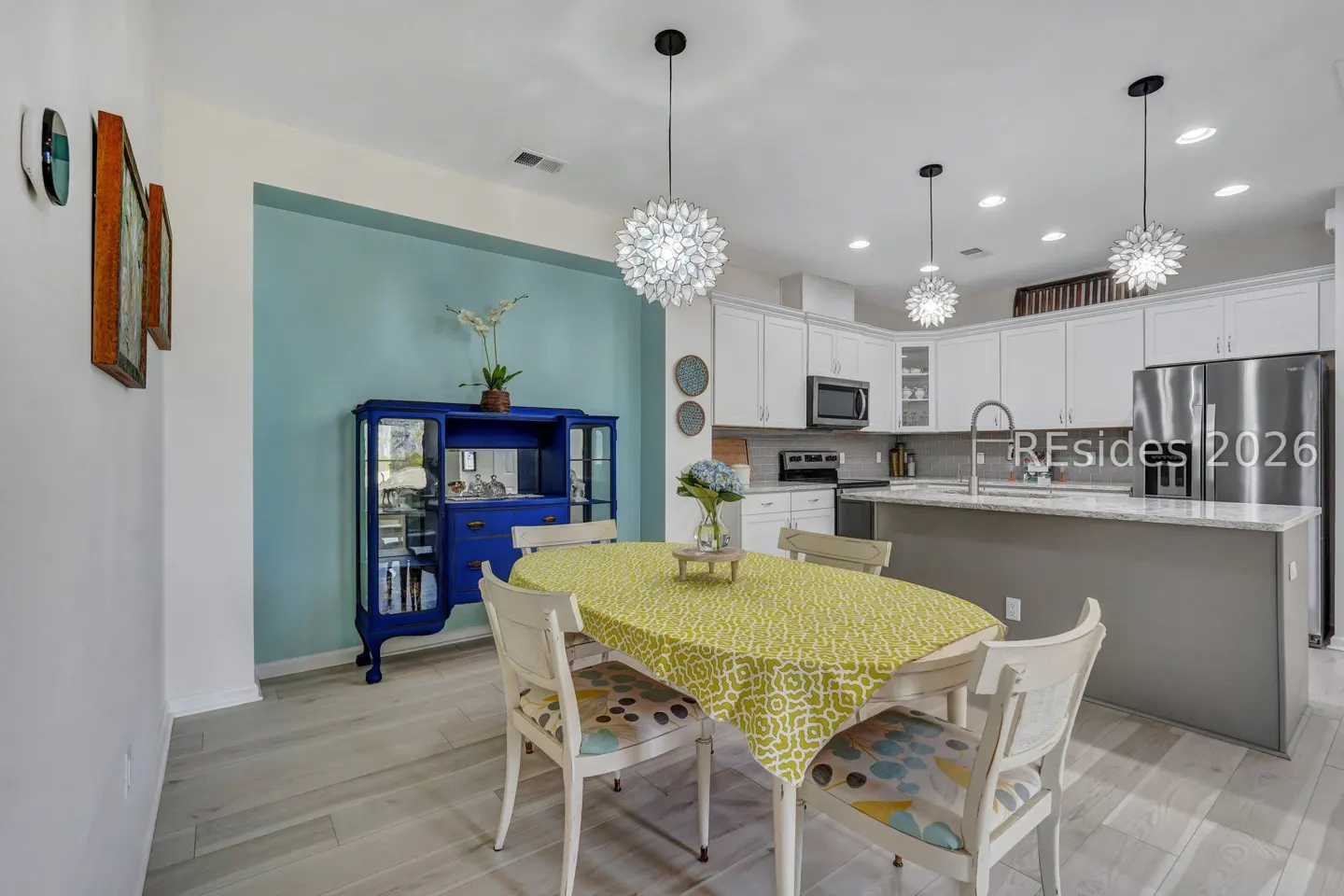 A bright, modern kitchen with a dining area. A blue hutch sits against a blue wall. A table with a yellow tablecloth is surrounded by white chairs.