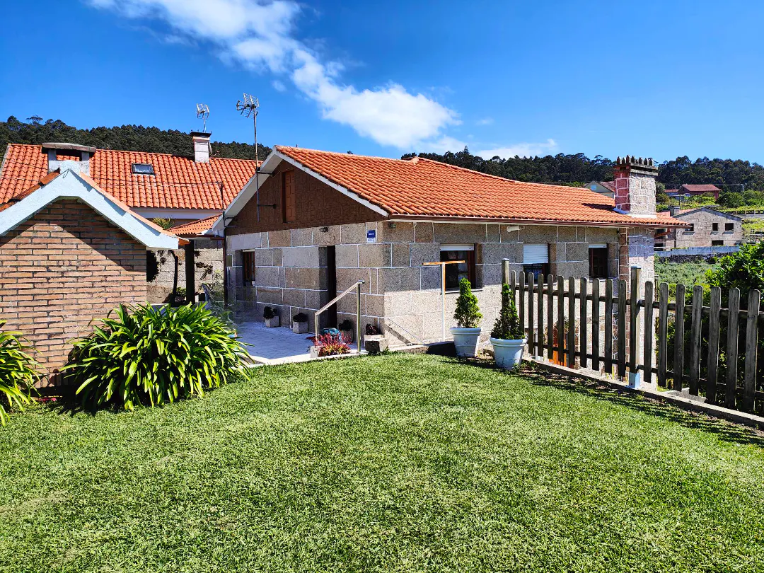 Exterior view of a stone house with a red tile roof, green lawn, and a wooden fence on a sunny day.