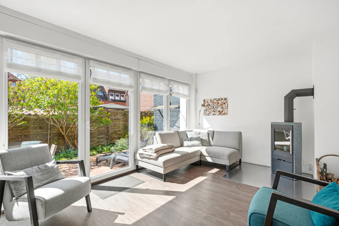 Bright living room with a gray sectional sofa, fireplace, and large windows overlooking a green backyard. Two armchairs are also visible.