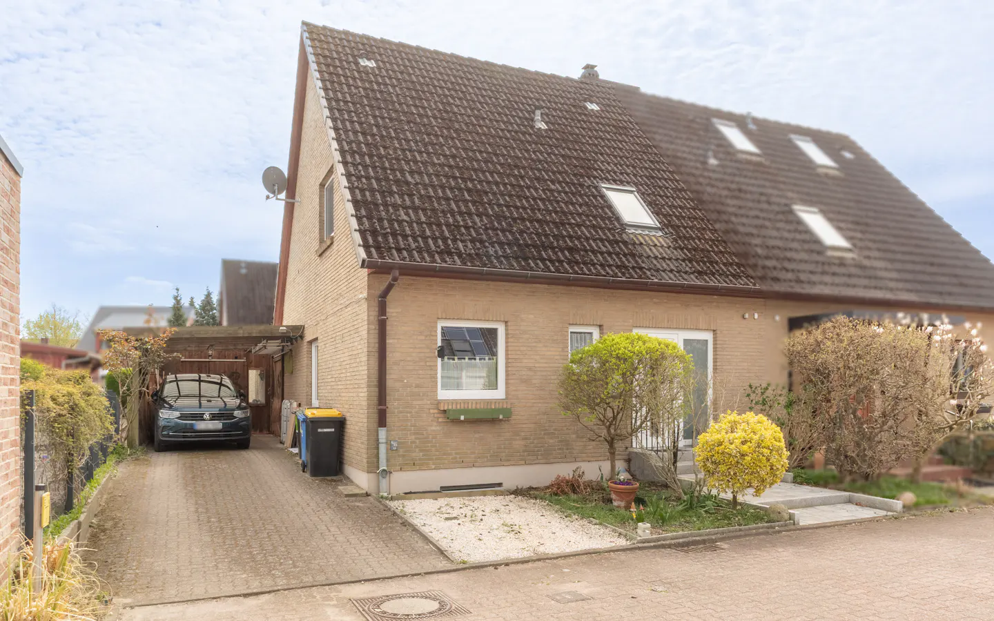 A tan brick house with a brown tiled roof, dormer windows, and a car parked in the driveway.