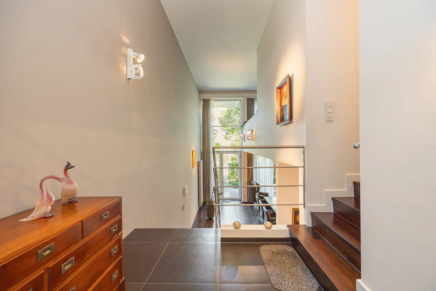 Entryway with dark tile floor, wooden dresser with flamingo statues, and stairs with stainless steel railing.