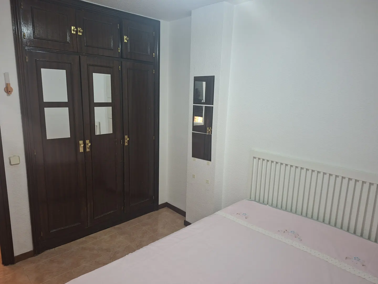 Bedroom with a pink bed, white headboard, and a dark wood wardrobe with gold handles.
