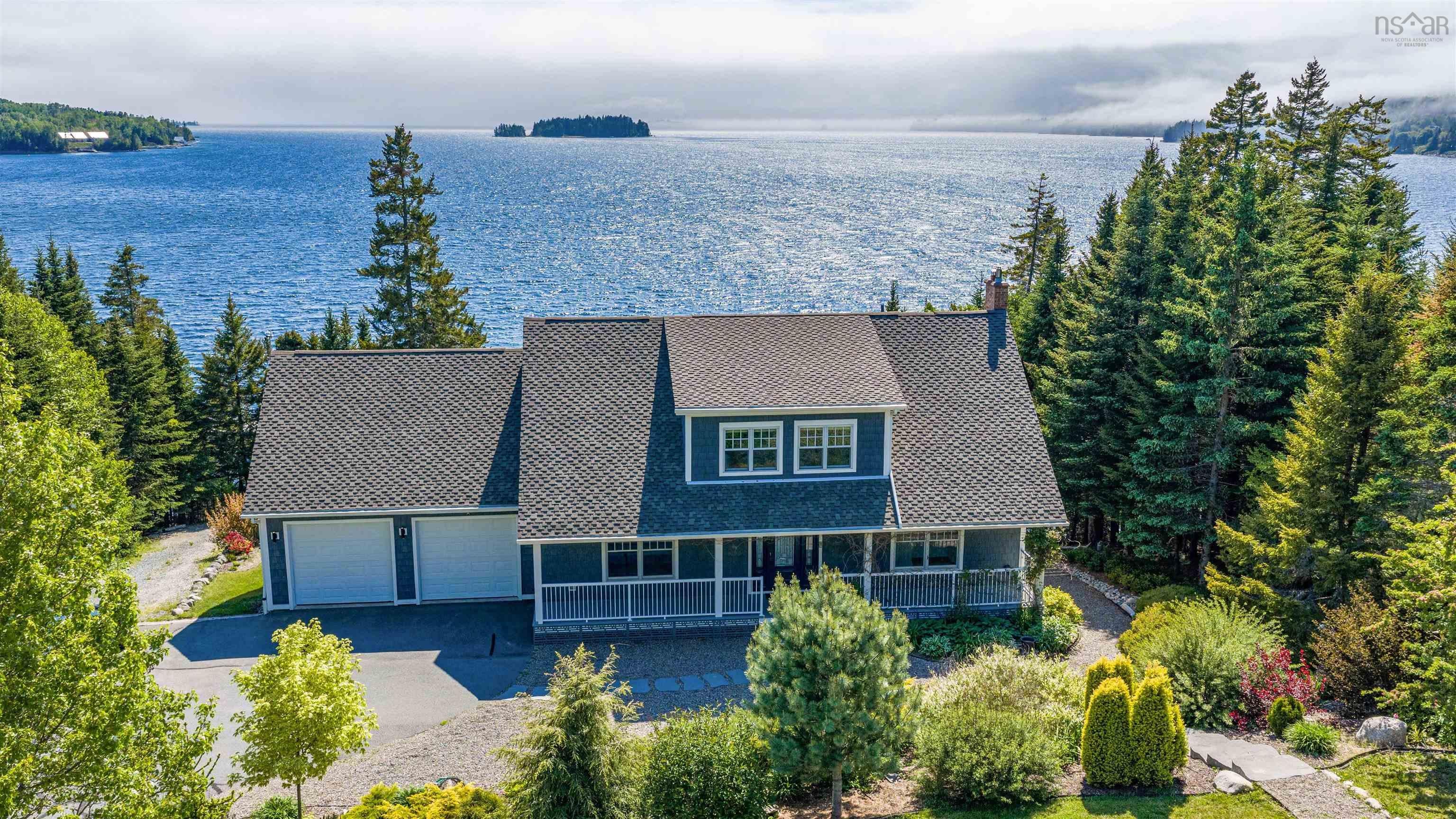 Aerial view of a blue two-story house with a double garage, surrounded by pine trees overlooking a calm, misty ocean bay. [AI-generated text]