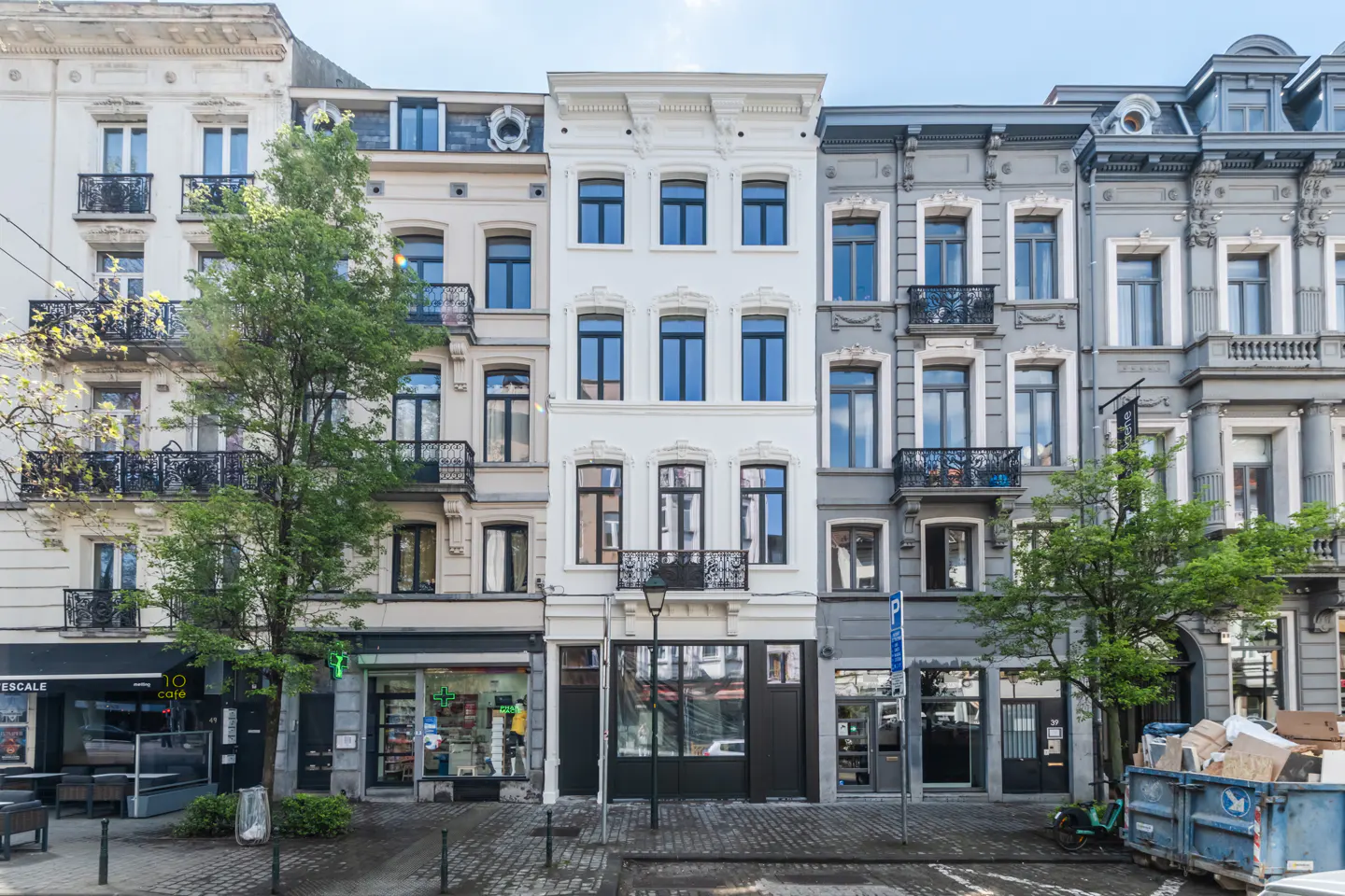 Row of multi-story buildings with shops on the ground floor. Buildings are white, beige, and gray with dark windows and small balconies.