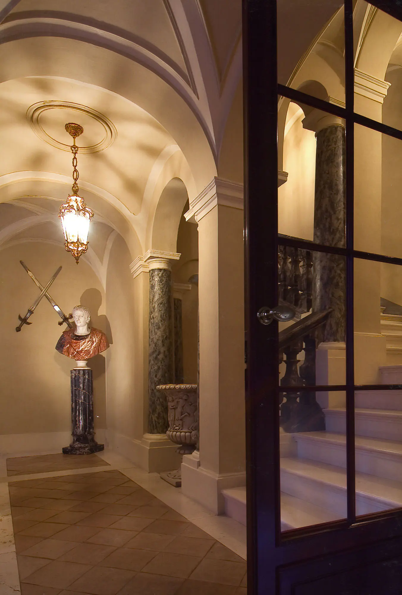 Elegant foyer with arched ceilings, marble columns, and a bust with crossed swords. Stairs are visible through an open door.