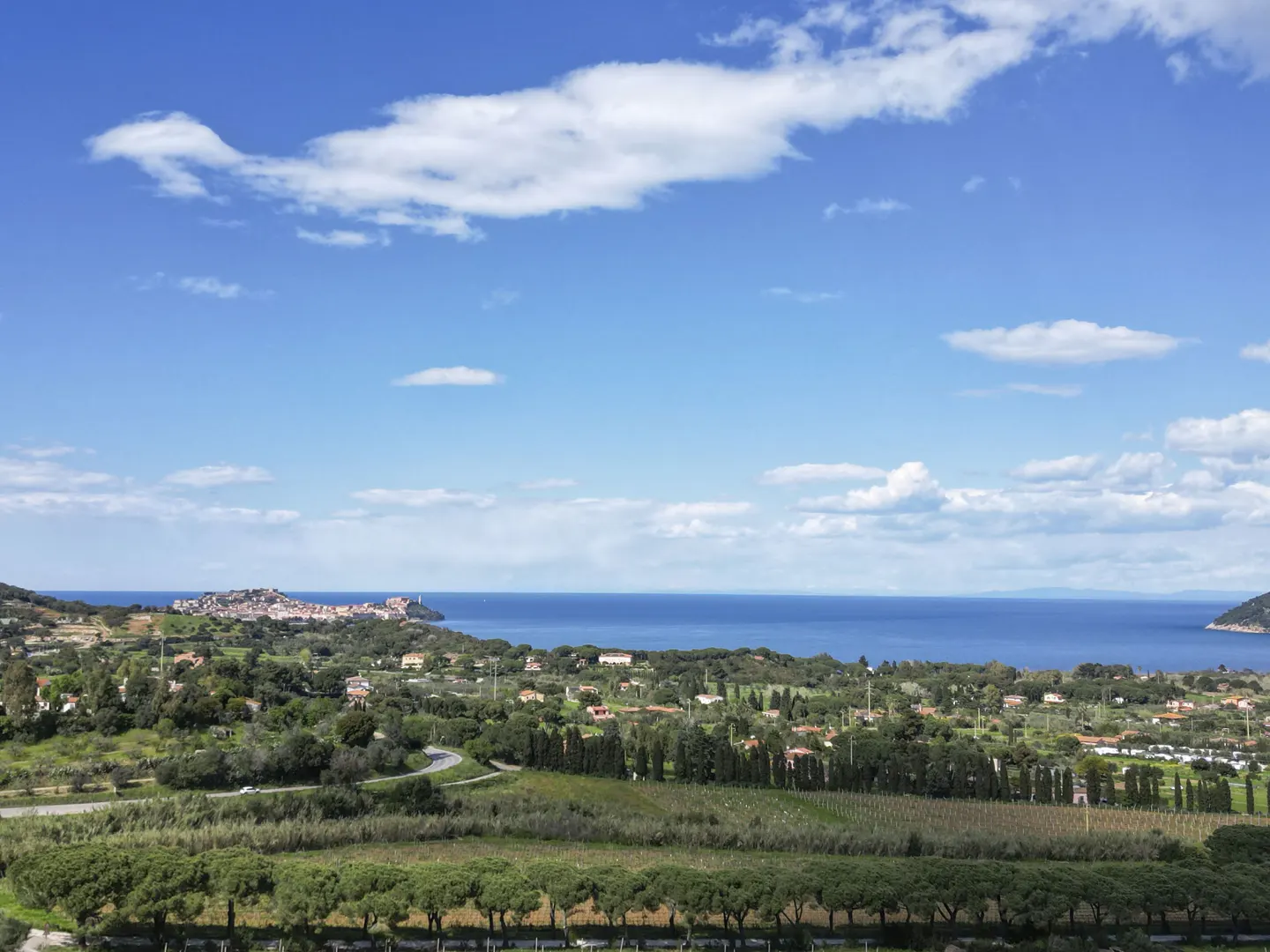 Scenic view of Elba Island with blue sky, clouds, and sea. Green hills, vineyards, and houses dot the landscape.