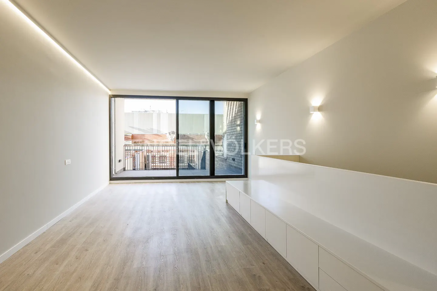 Bright, modern room with light wood floors, white walls, and a sliding glass door to a balcony. Built-in white storage units line the wall.