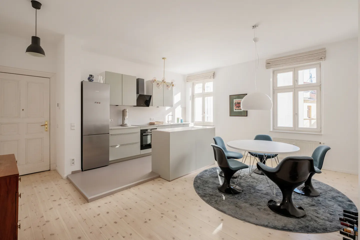 Bright, modern kitchen with light wood floors, gray cabinets, stainless steel fridge, and a white dining table with blue chairs.