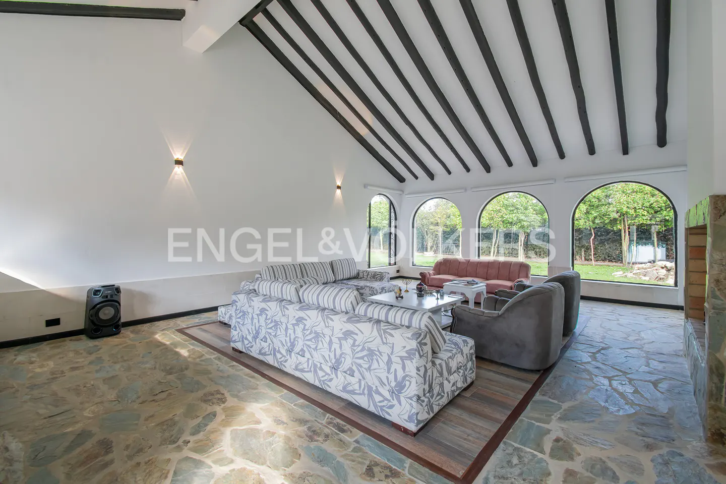 Bright living room with stone floors, white walls, and dark wood beams. Large arched windows overlook a green lawn. Sofas and chairs provide seating.