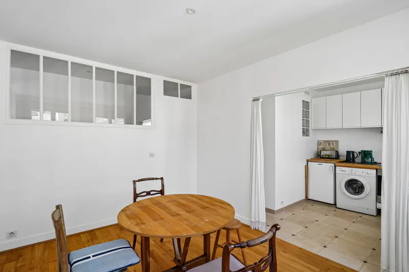Bright dining area with wood floors, a round table, and chairs. A kitchen with white cabinets and appliances is visible through a doorway.