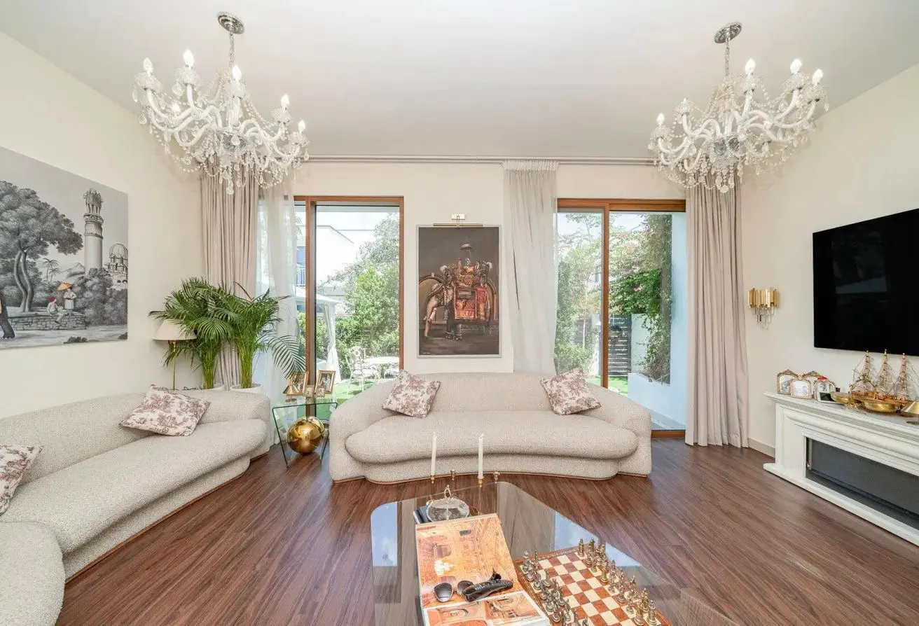 Living room with two white sofas, a glass coffee table, and two crystal chandeliers. Large windows look out onto a green yard.