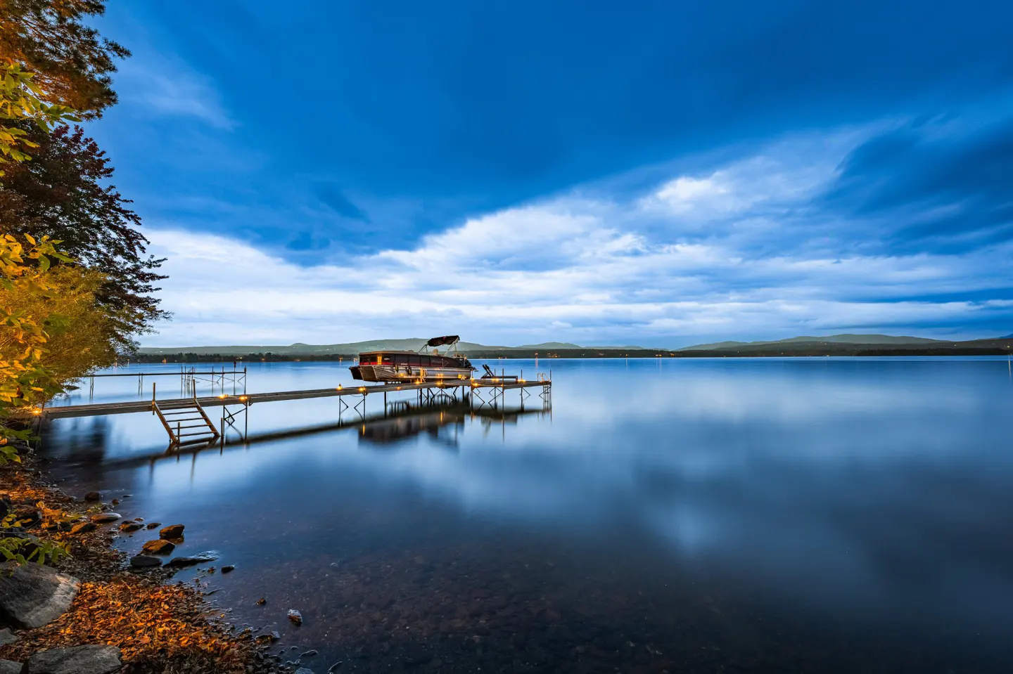 A serene lake view with a boat docked on a wooden pier under a cloudy blue sky.