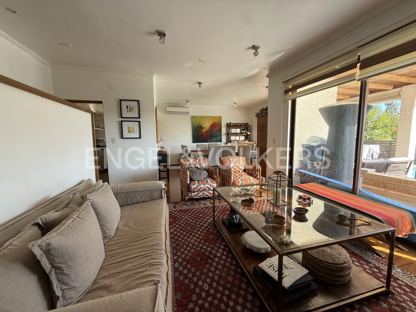 Bright living room with a beige sofa, patterned armchairs, and a glass coffee table on a red rug. Large windows overlook the outdoor area.