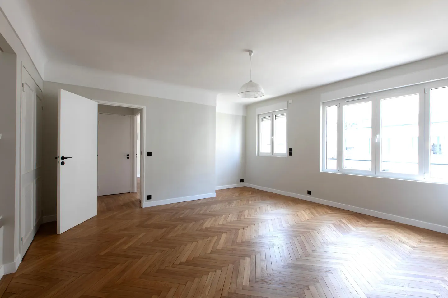 Empty room with light gray walls, herringbone wood floors, white doors, and large windows letting in natural light.
