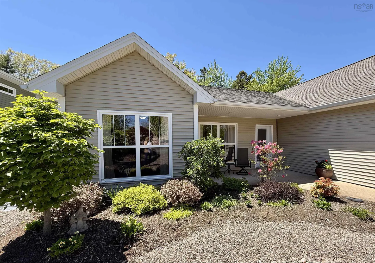 Single-story beige house with a covered porch, gravel landscaping, and flowering shrubs under a clear blue sky.