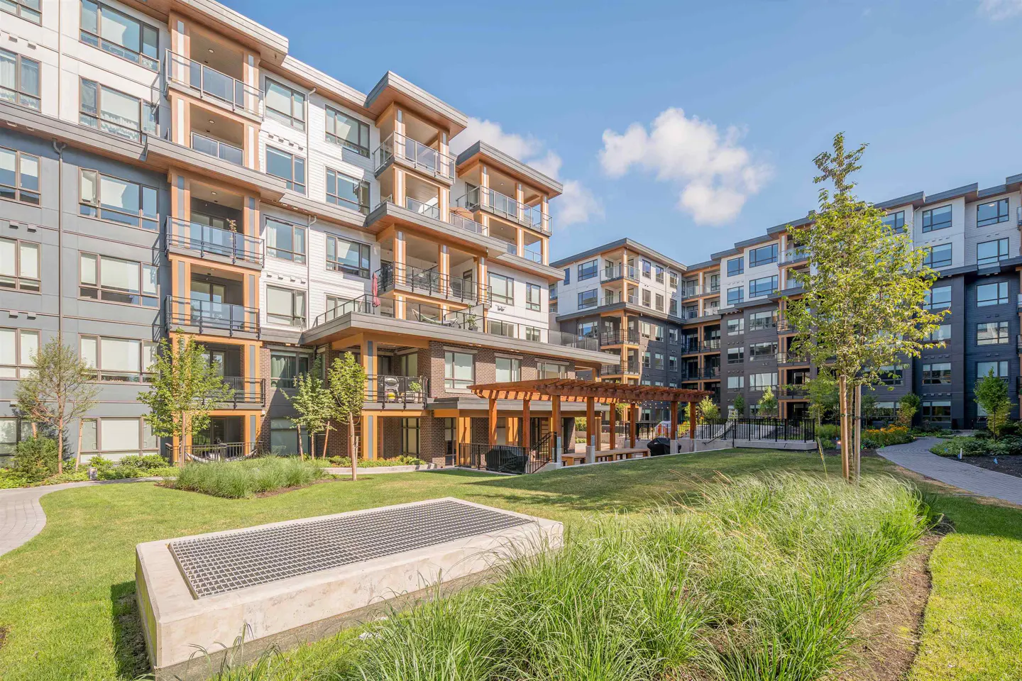 Exterior view of a modern apartment complex with a central green space. Balconies, wood accents, and a pergola enhance the building's design.