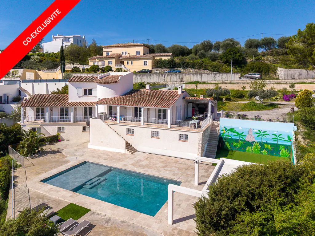 Aerial view of a white villa with a tiled roof, a swimming pool, and a mural-painted wall in a sunny, landscaped garden.
