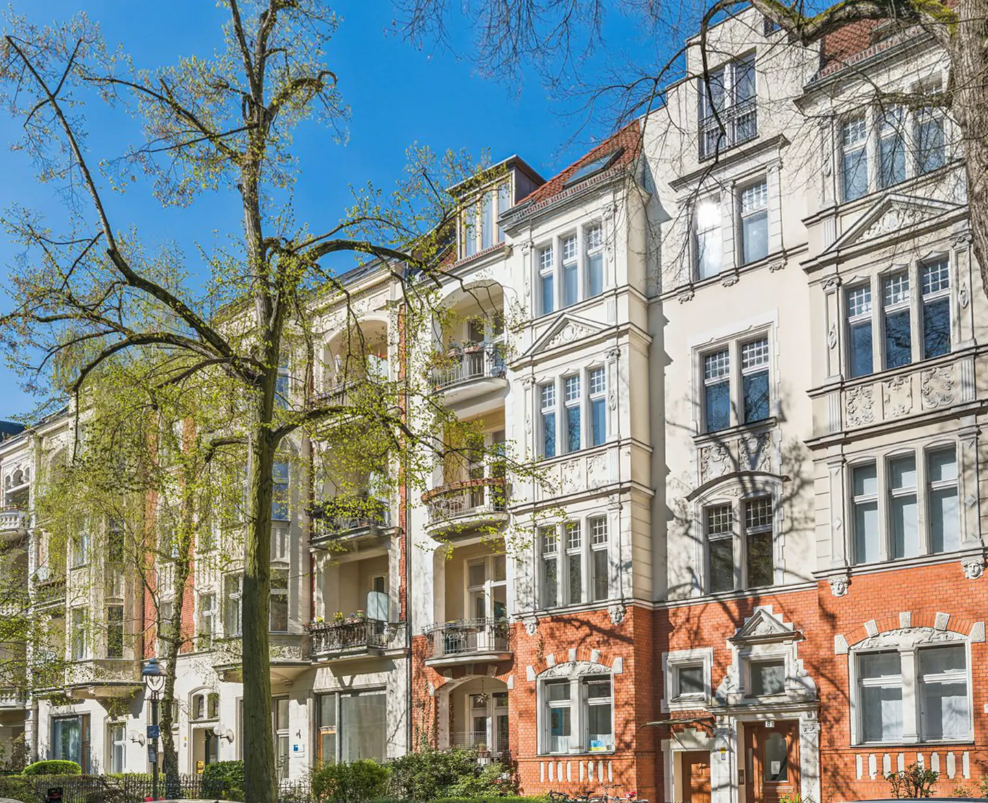 Row of multi-story apartments with white facades and red brick accents under a blue sky, framed by trees.