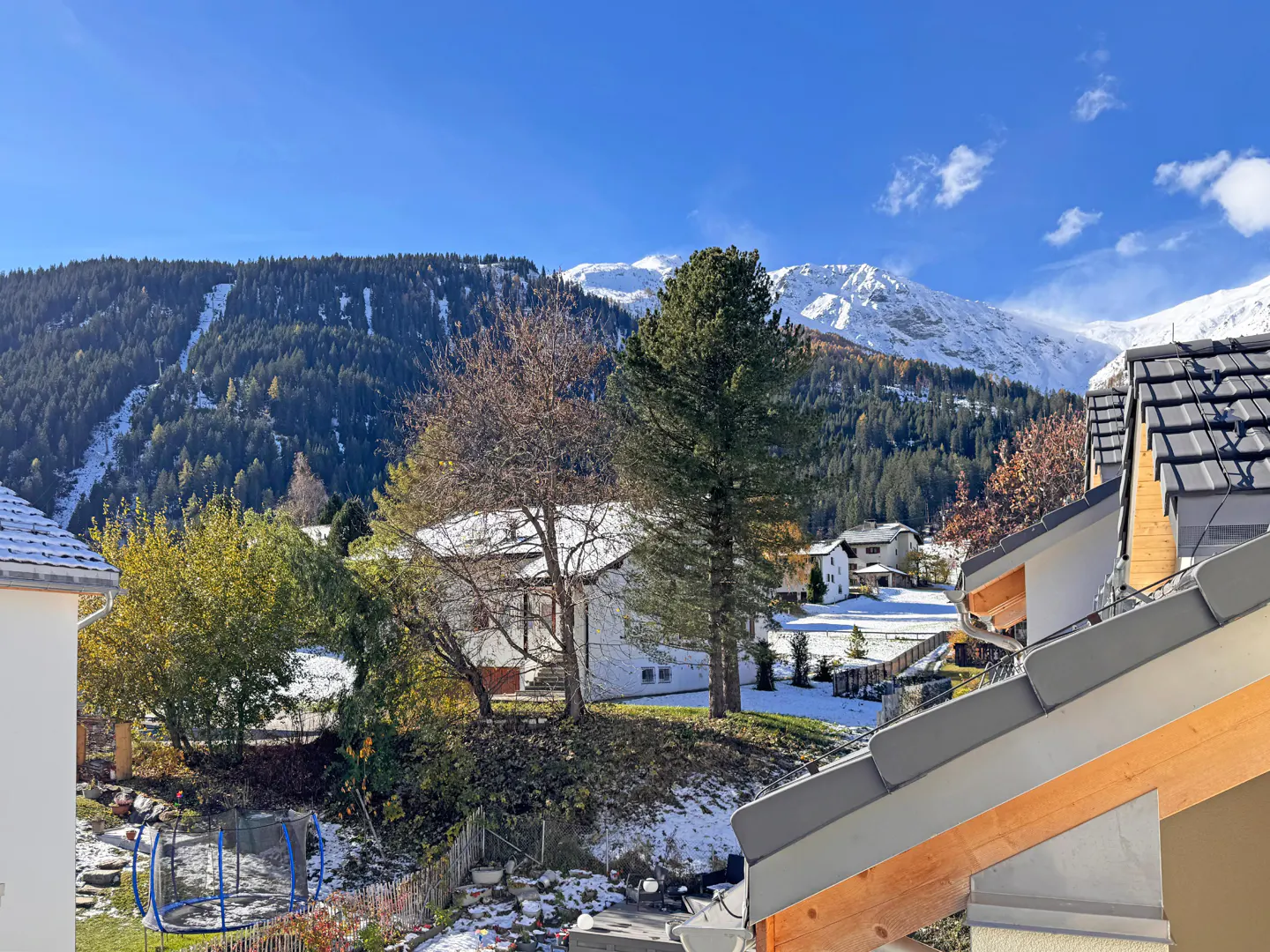 Scenic view of a Swiss village with snow-capped mountains, trees, and rooftops under a clear blue sky.