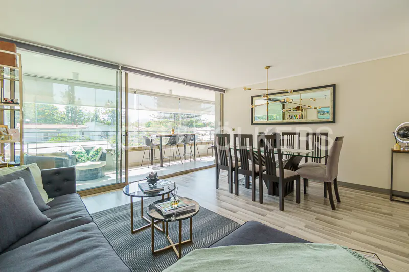 Bright living room with gray sofa, glass coffee tables, and dining area with glass table and black chairs. Balcony with seating.