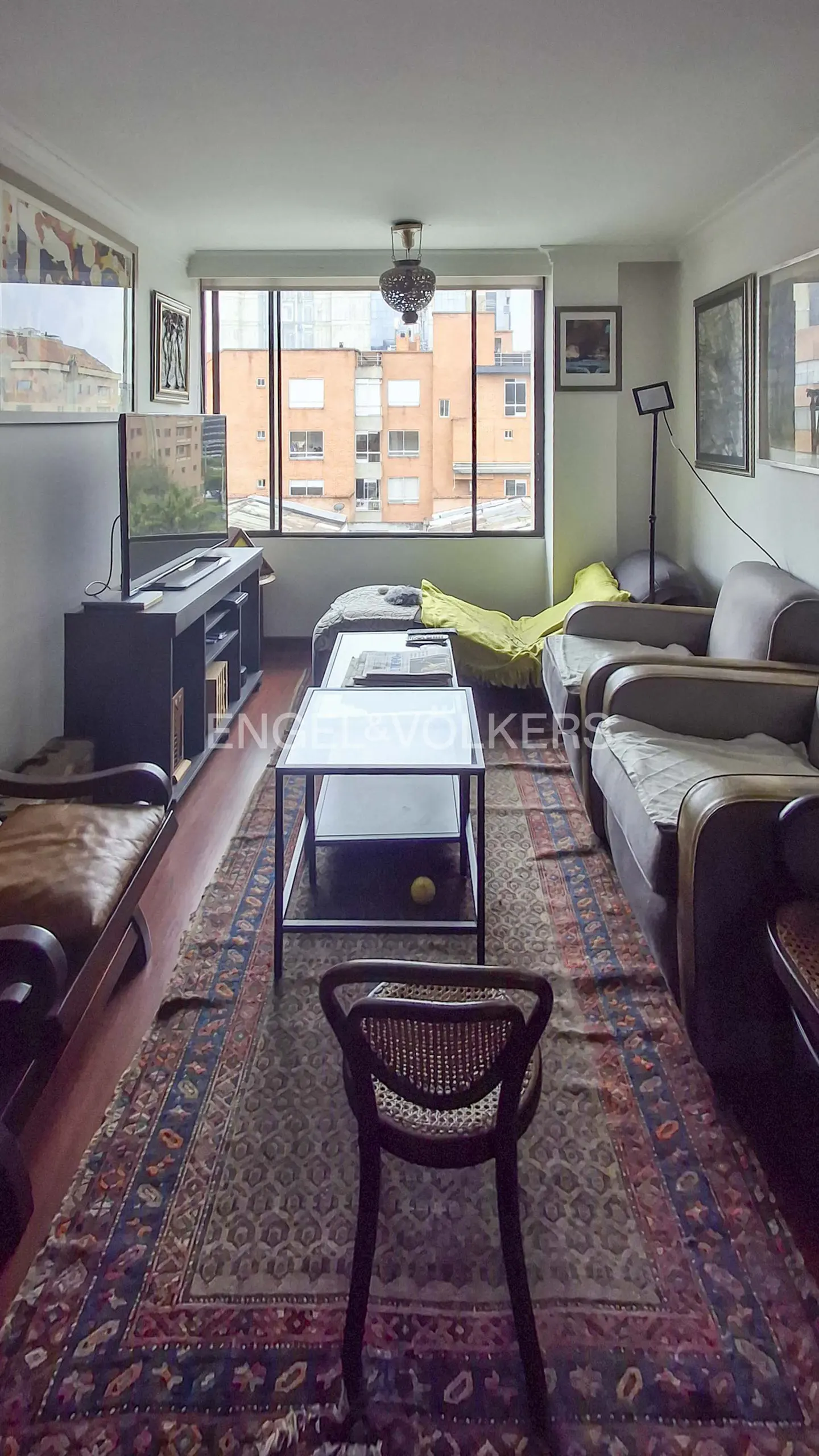 Living room with a large window, sofas, a TV, and a patterned rug. A chair faces the viewer.