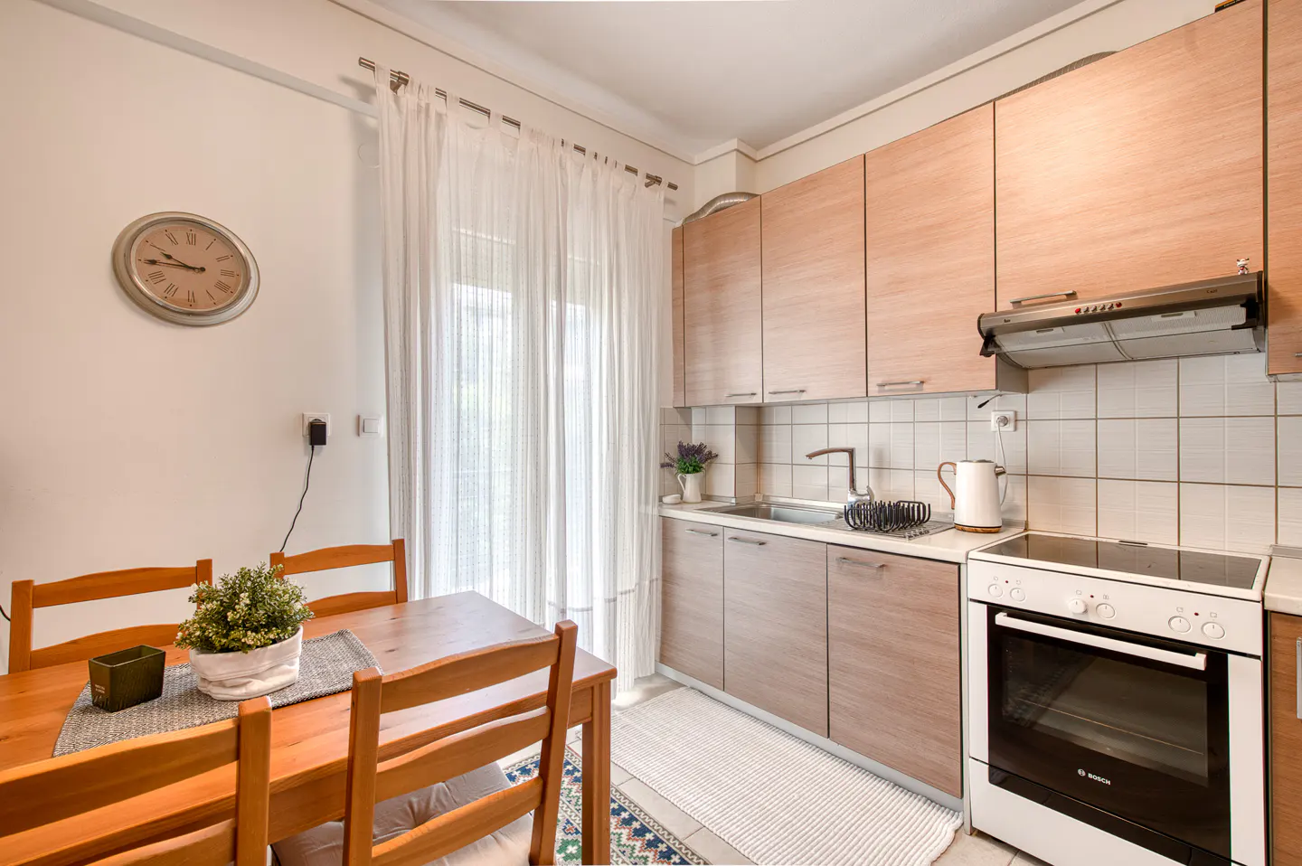 A bright kitchen and dining area with wood cabinets, a white stove, and a wooden table with chairs. A clock hangs on the wall.