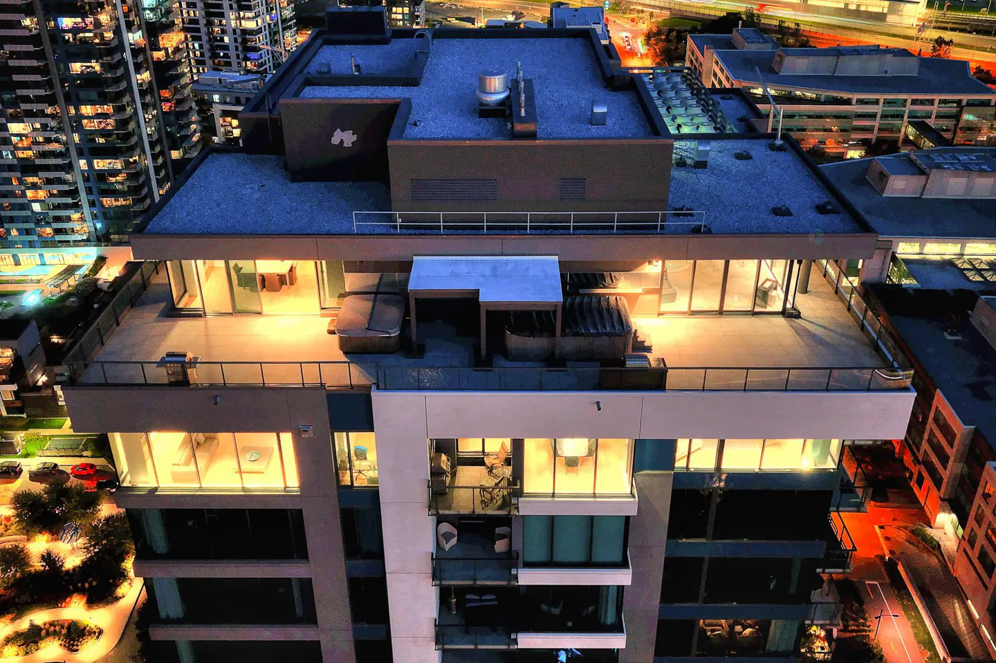 Aerial view of a modern high-rise building at night, featuring a rooftop terrace with a hot tub and city lights in the background.