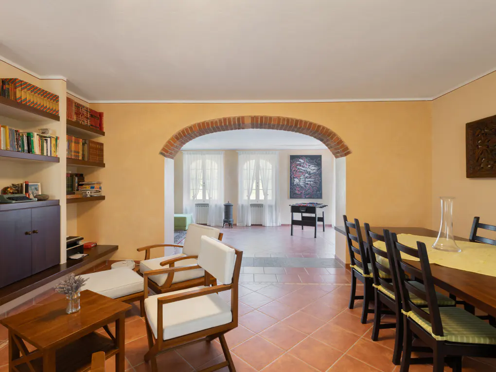 Living room with terracotta tile floor, arched doorway, bookshelves, and dining table with chairs. Walls are light orange.