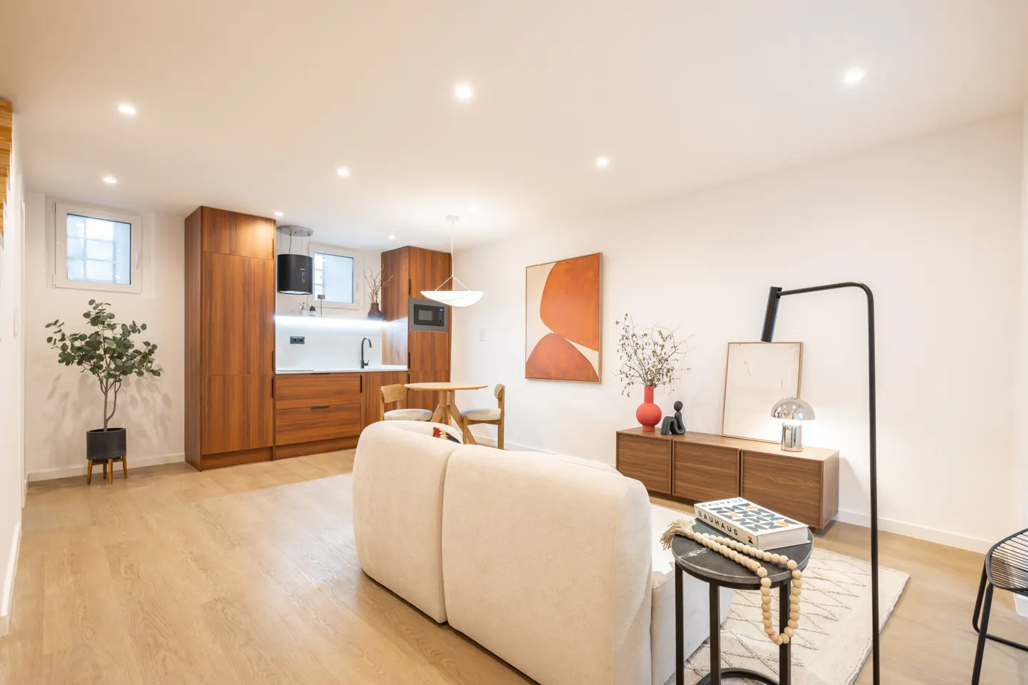 Bright, open-concept apartment with wood floors, white walls, and recessed lighting. A cream sofa faces a wood cabinet with art. A small kitchen is in the background.
