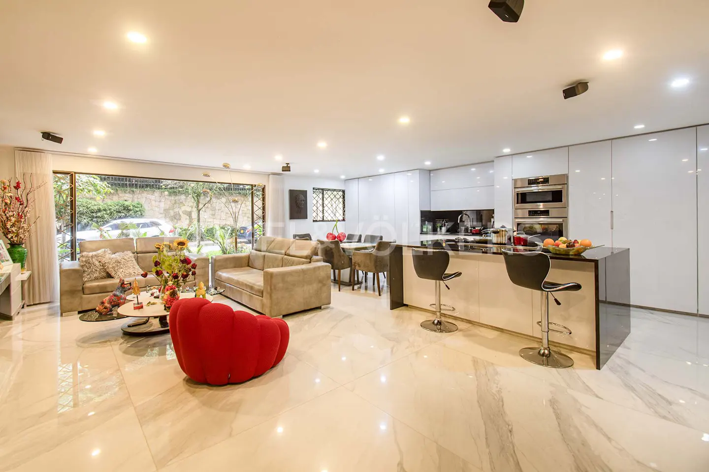 Open-concept living space with tan sofas, red chair, and kitchen island with stools. Large windows offer a view of greenery.