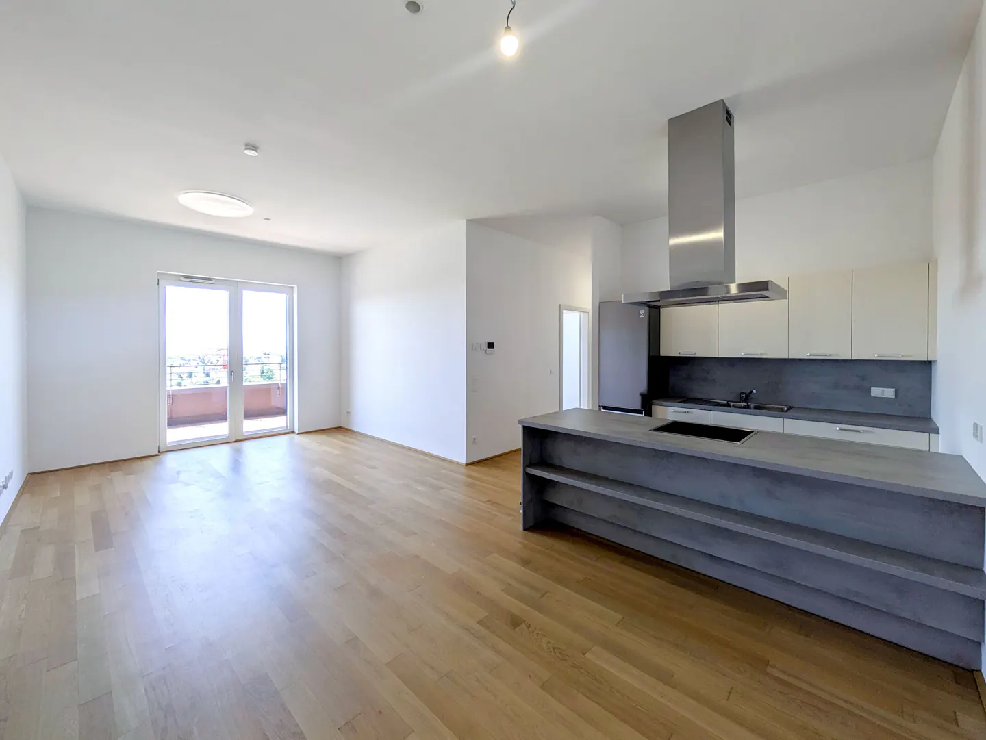 Modern open-plan kitchen with light wood floors, white cabinets, grey island, and glass doors leading to a balcony.