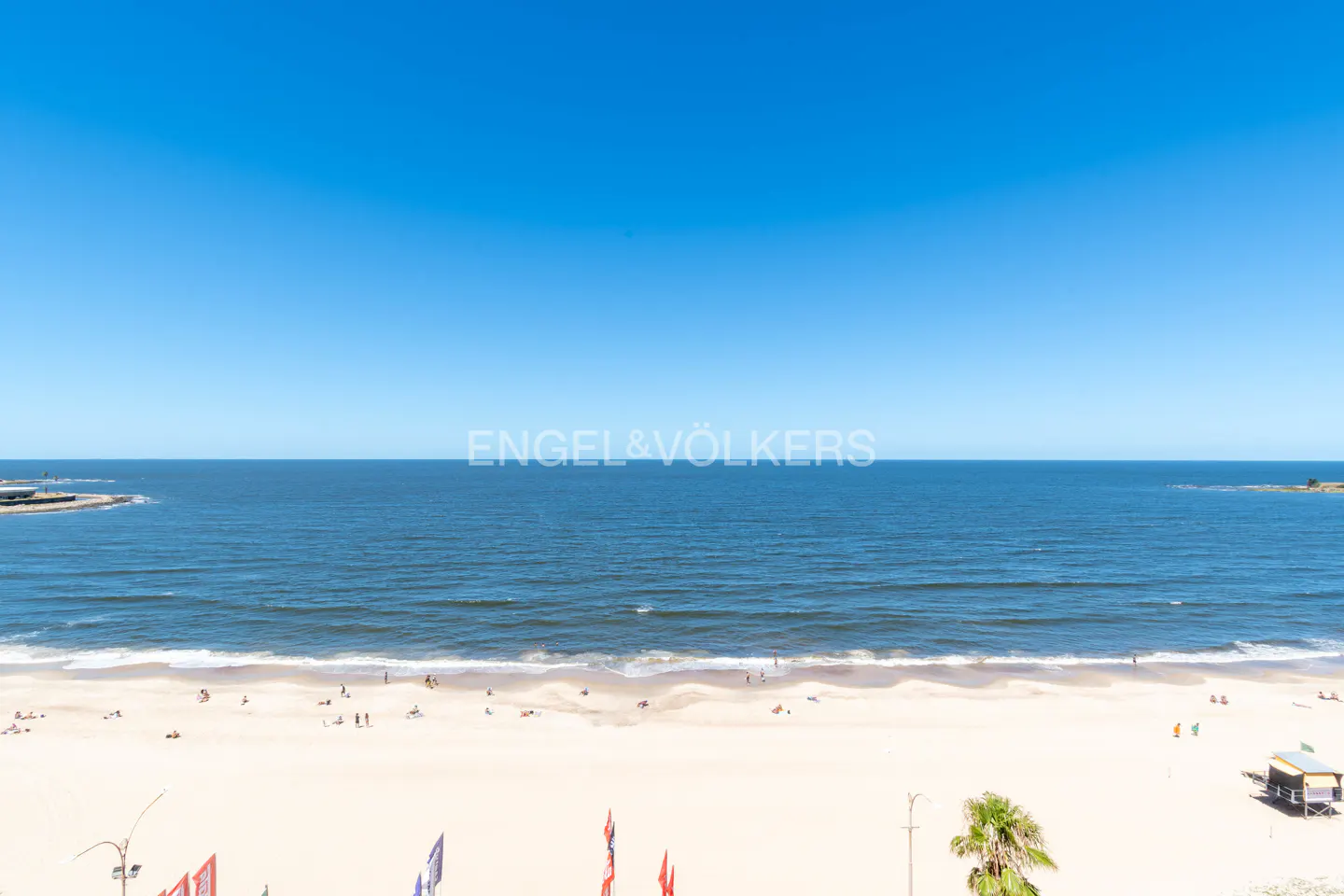 Beach view with blue sky, ocean, and sandy shore. People are scattered on the beach, and a lifeguard station is visible.