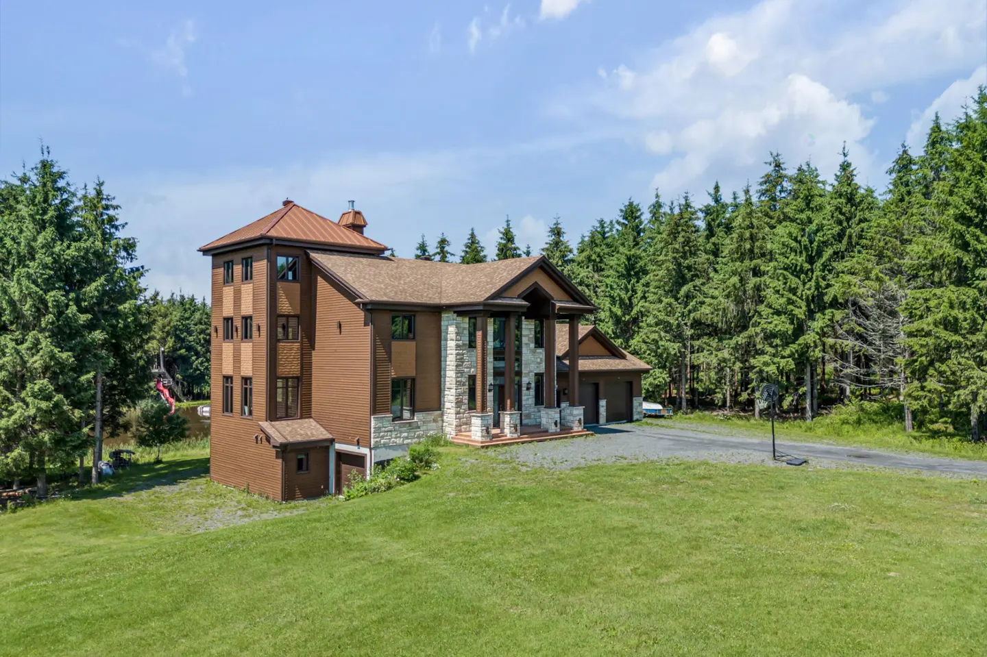 Three-story brown house with stone accents, a multi-level roof, and a garage, set on a grassy lot near a pine forest.
