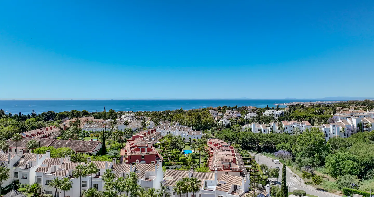 Aerial view of a coastal community with white and red buildings, green trees, and a blue ocean under a clear blue sky.