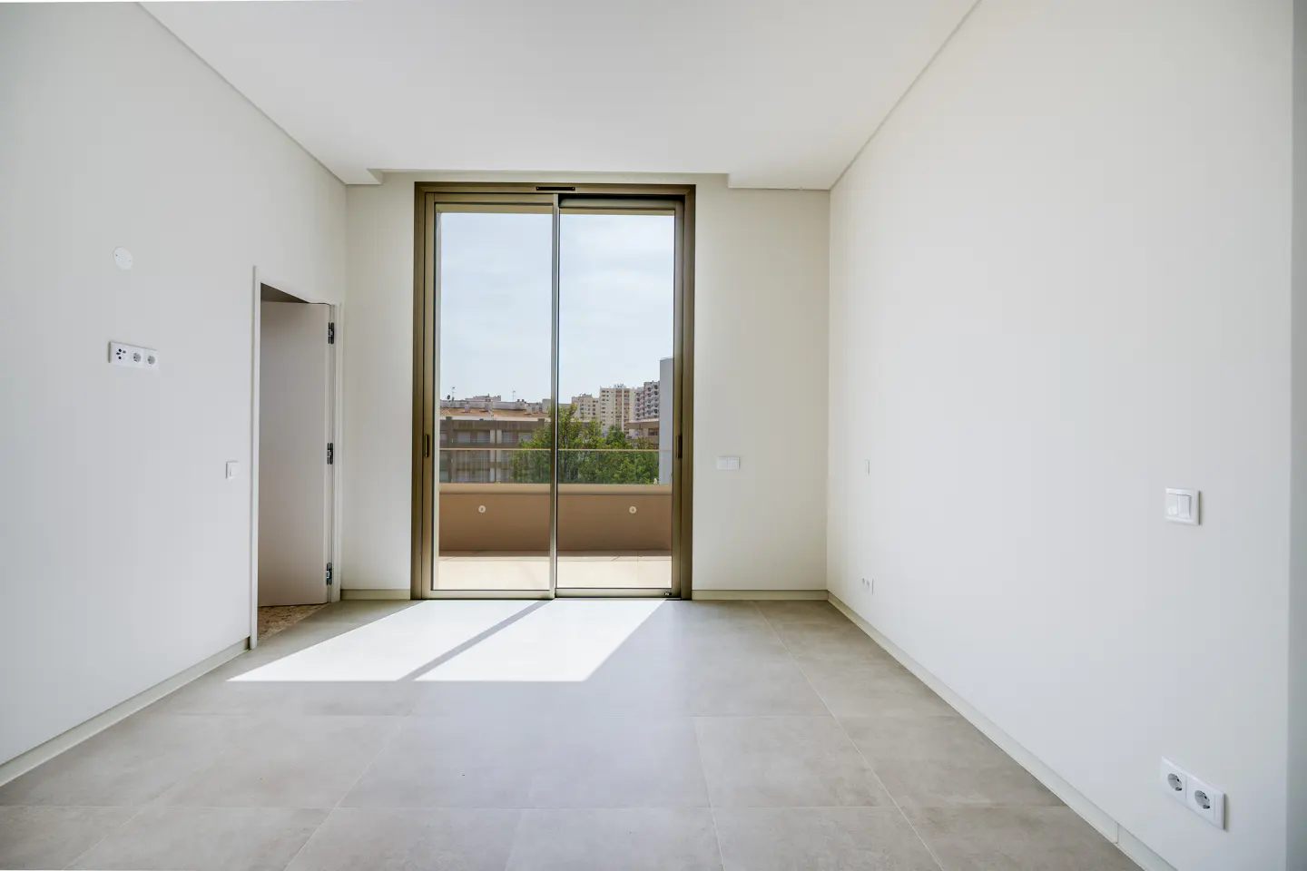 Bright, empty room with gray tile floor, white walls, and sliding glass doors to a balcony with city views.
