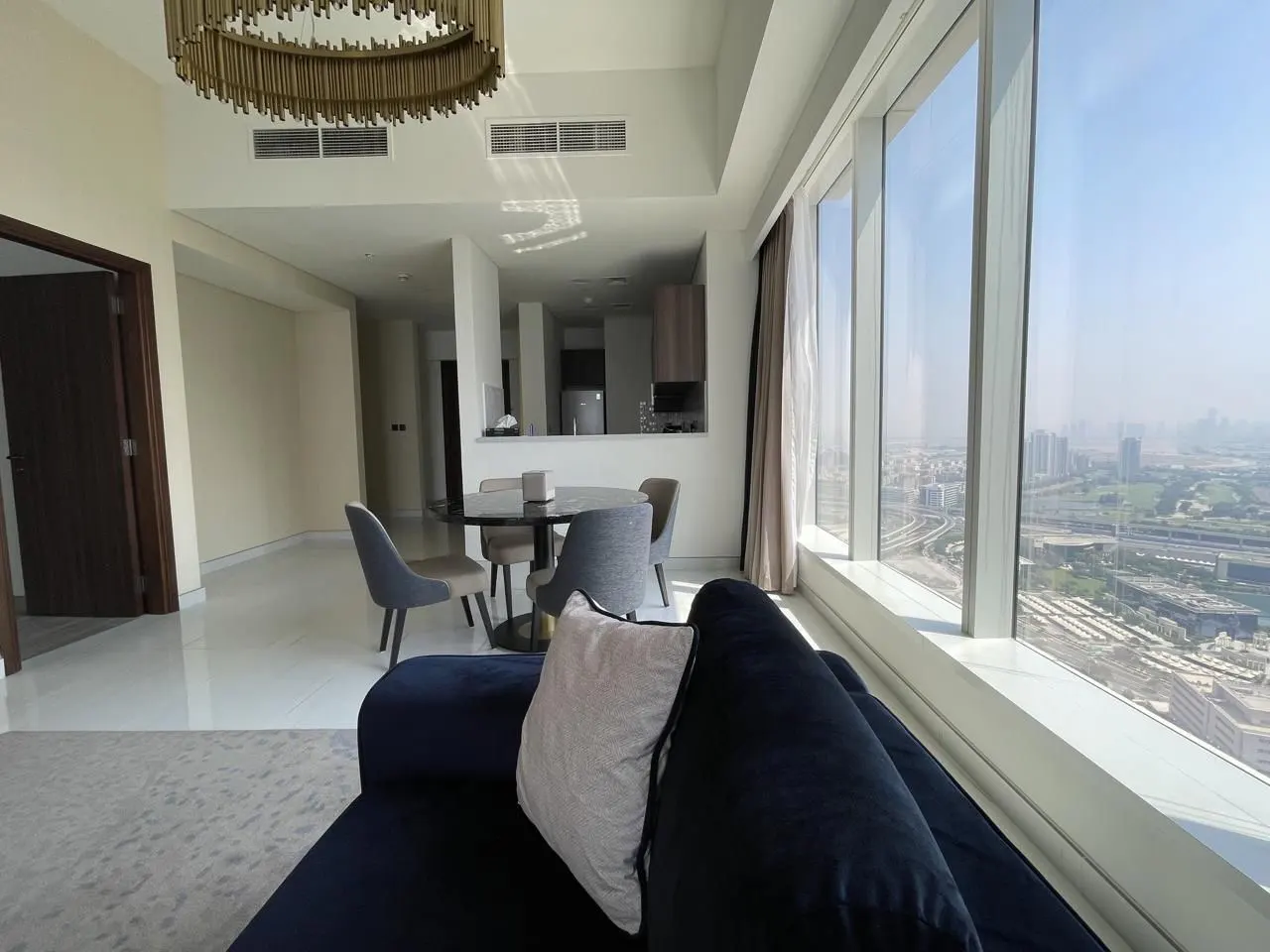 Bright apartment interior with a blue sofa, round dining table, and floor-to-ceiling windows overlooking a cityscape.