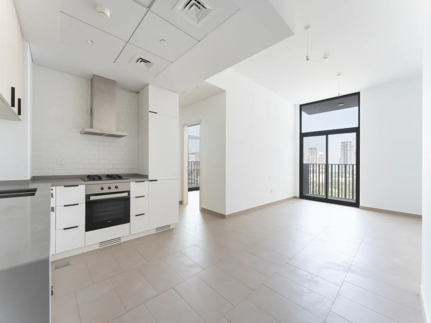 Bright, modern apartment interior with white walls, tile floors, and a kitchen with stainless steel appliances. A large window offers a city view.