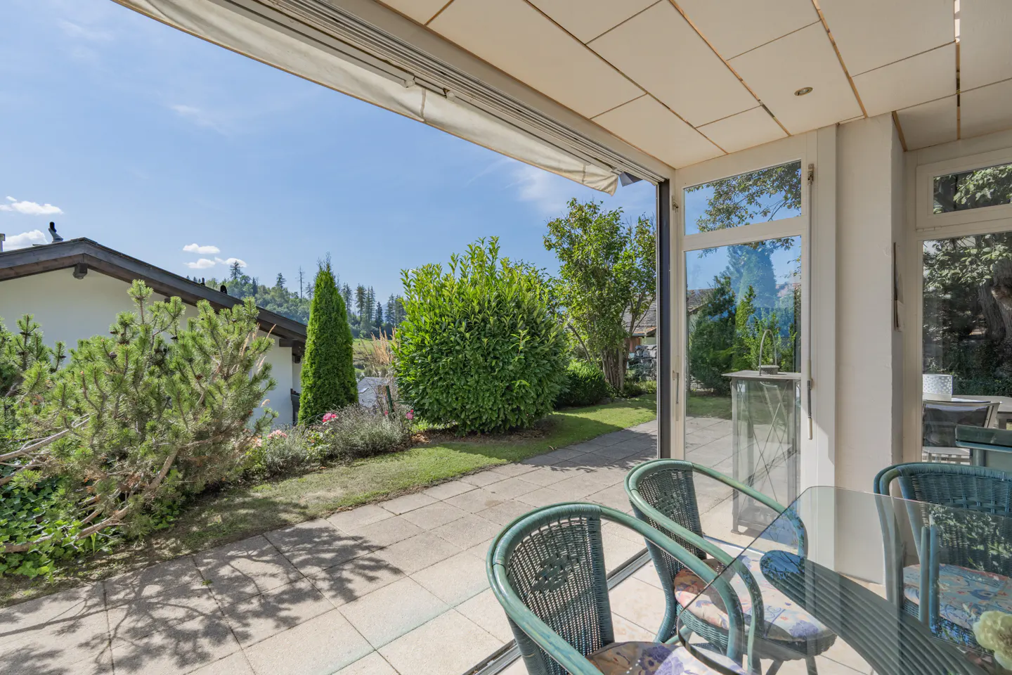 Patio view of a garden with green chairs and a glass table. Outside is a lawn, trees, and a house under a blue sky.