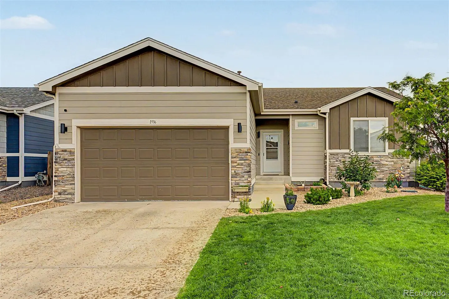 Single-story suburban home with tan siding, stone accents, a two-car garage, and a green front lawn under a blue sky.